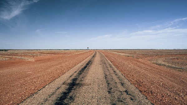 A desert road, somewhere in the outback. Something can be just made out in the long distance, but it is not clear what it is.